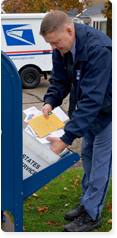A male letter carrier picking up mail from an outdoor post box. He is dressed in a postal uniform with a light blue shirt, blue slacks and a navy jacket.