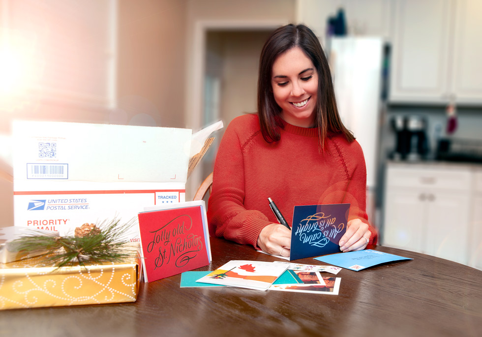 Woman at her kitchen table writing holiday letters with USPS packaging on the table