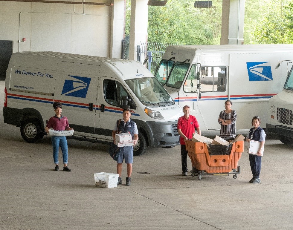 Postal employees infornt of vehicles processing mail and packages.