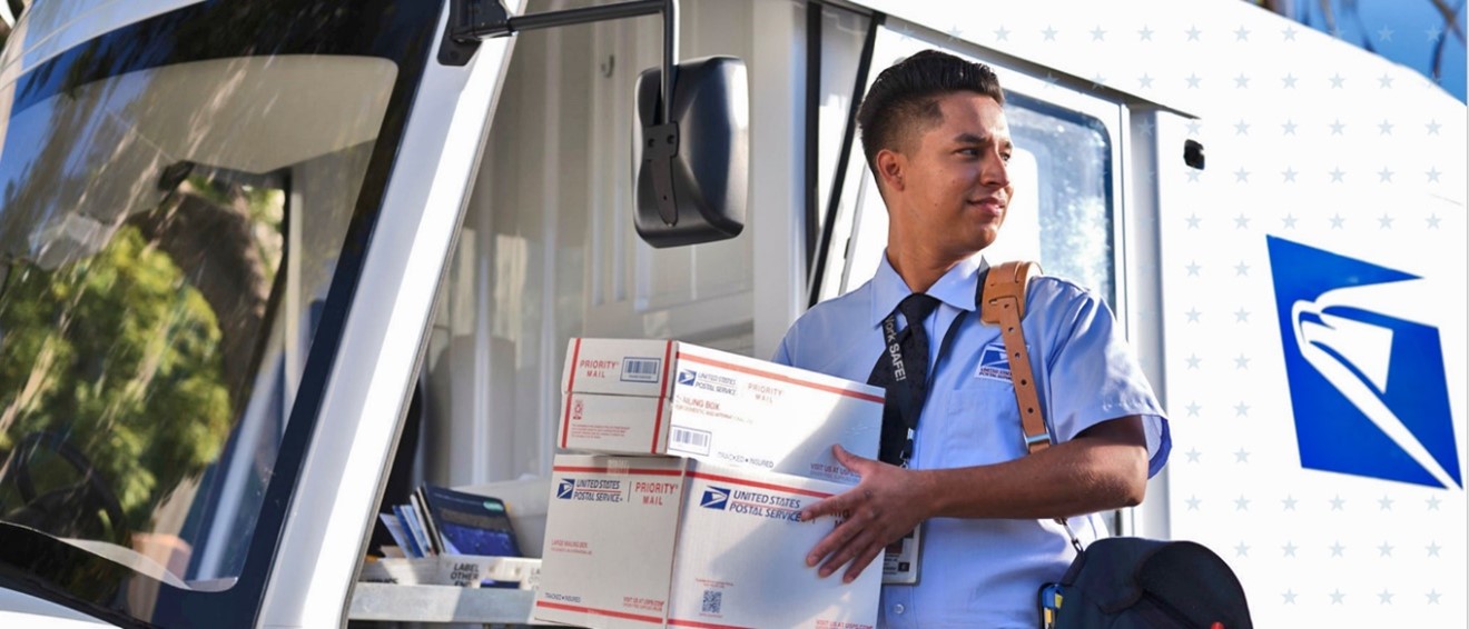 USPS Letter Carrier holding Priority mail packages in front of mail vehicle.