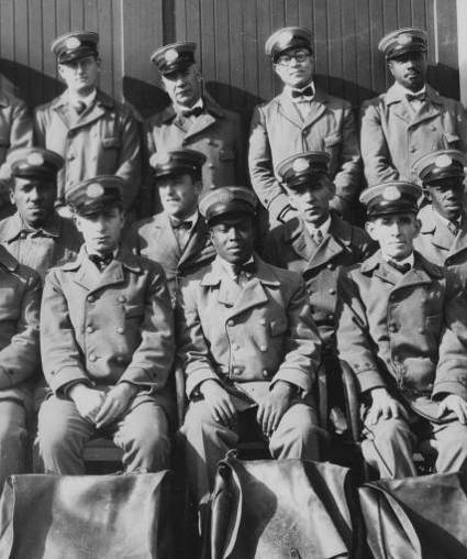 Detail of a photograph of a group of uniformed letter carriers, both black and white, posing with mail satchels in 1926.