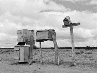 This photograph shows a group of four rural mailboxes on wooden posts in the foreground, and miles of empty desert in the background.