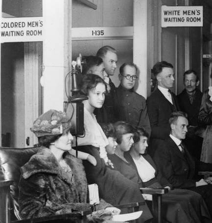 Photograph showing white federal workers in a waiting room at the U.S. Public Health Service dispensary in Washington, D.C., circa 1918.  A sign overhead reads 'White Men's Waiting Room.'  An adjacent sign overhead reads 'Colored Men's Waiting Room.'